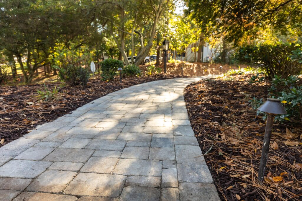 Curved stone pathway in a garden with mulched beds and green shrubs under sunlight, featuring a small path light on the side.