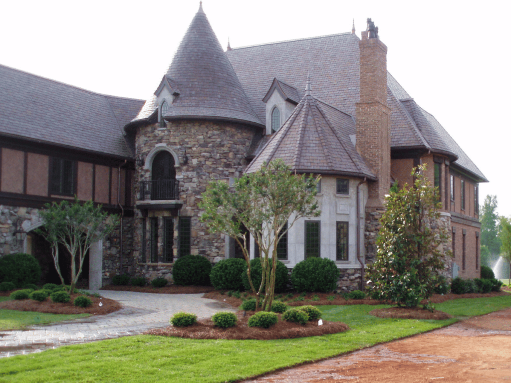 Large stone and brick house with a turret and a front yard landscaped with hardy plants and shrubs