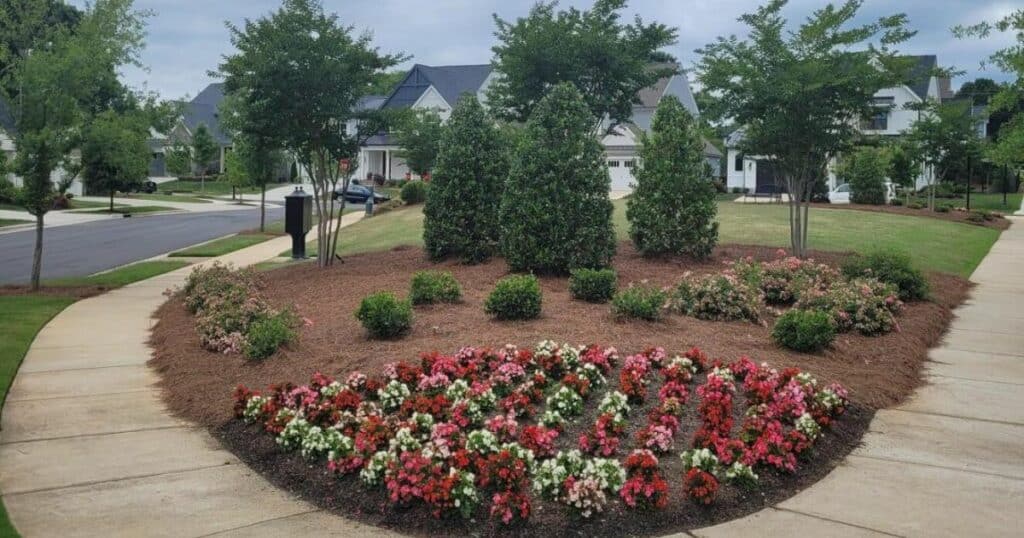 A neighborhood intersection features a landscaped roundabout with rows of vibrant flowers, bushes, and trees, bordered by mulch and surrounded by sidewalks and residential houses.