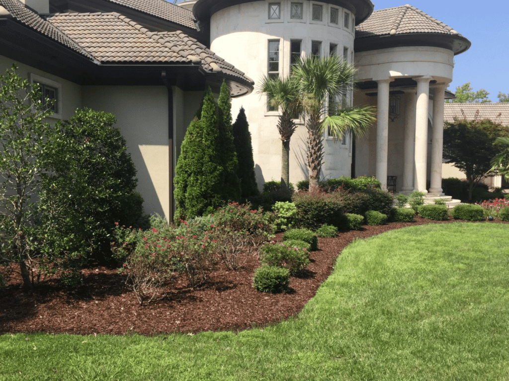 A well-maintained garden completed with landscape design plans with shrubs, a small palm tree, and mulch complements the large, elegant house with a cylindrical tower and columned entrance, showcasing meticulous landscape design plans.