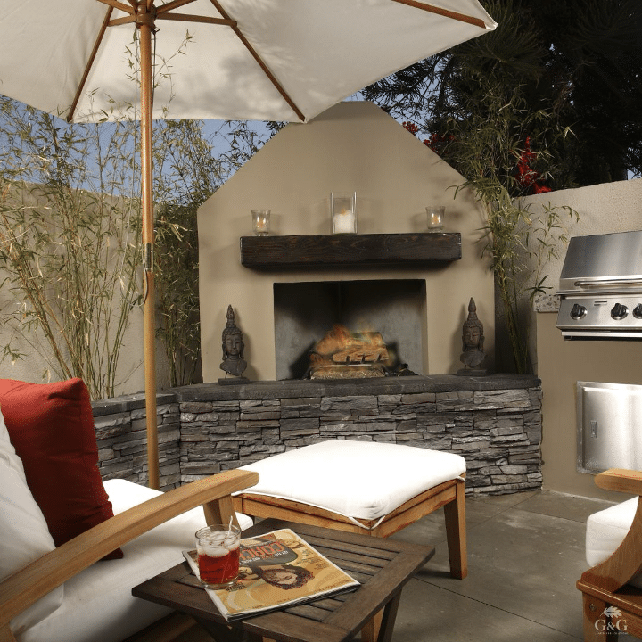 Outdoor patio area with a large umbrella, comfortable seating, a small table with drinks and a magazine, a stone fireplace, and a nearby barbecue grill against a background of plants and trees.