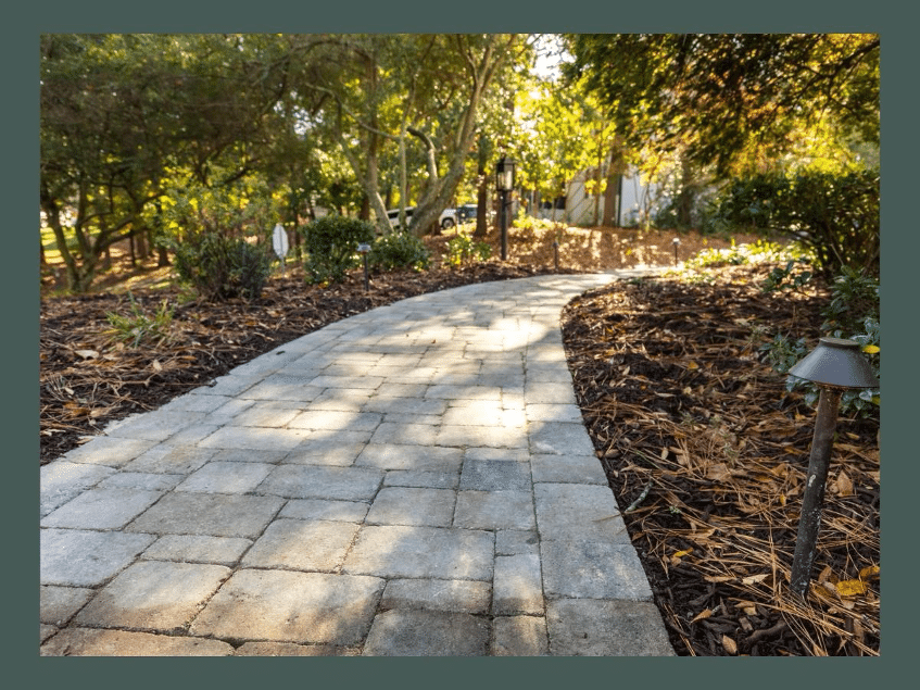 A stone-paved pathway curves through a park-like area, with trees and foliage on both sides and a garden light on the right.