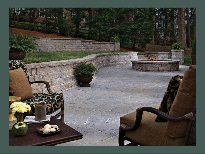 A stone patio with lounge chairs and a low table, featuring a raised brick fire pit and surrounded by a landscaped garden with a stone retaining wall and trees in the background.