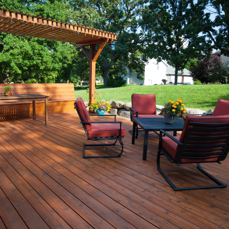 A wooden deck with a pergola, red-cushioned chairs, a table, and flower pots. A grassy backyard with trees is visible in the background.