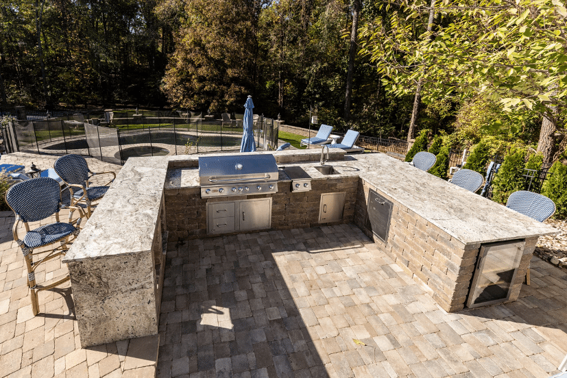 Outdoor kitchen setup with granite countertops, built-in grill, sink, and storage cabinets on a patio. Outdoor seating and a fenced pool area are visible in the background.