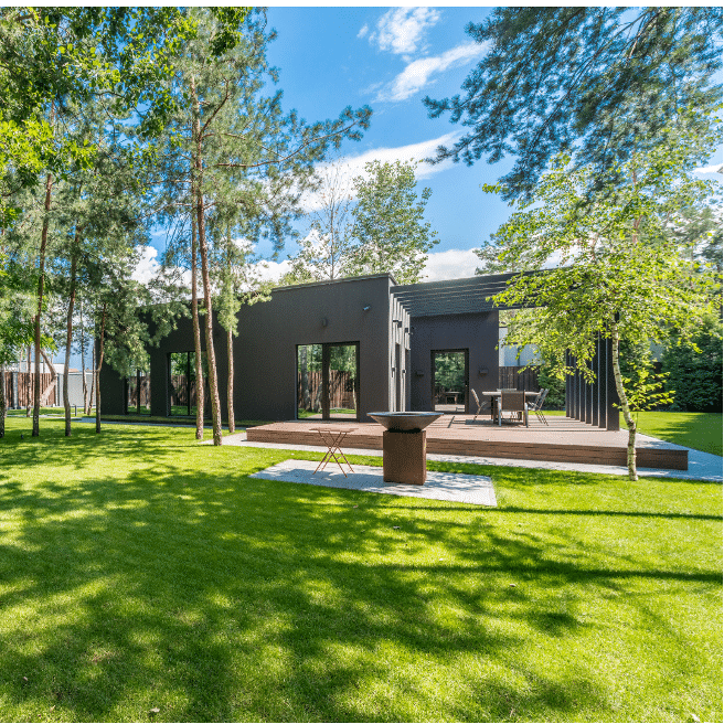 Modern one-story black house with large windows, surrounded by trees and a green lawn. It features an outdoor seating area and a fire pit on a wooden deck. Clear blue sky above.