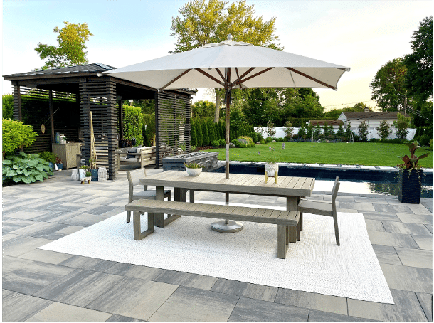 A modern outdoor patio features a large wooden table with benches under a white umbrella. In the background, there is a shaded seating area and a pool surrounded by greenery.