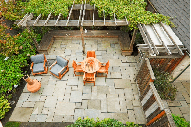 Aerial view of a stone-paved patio with a round wooden table, four chairs, two cushioned lounge chairs, a chimney-style fireplace, and a wooden pergola covered in greenery.