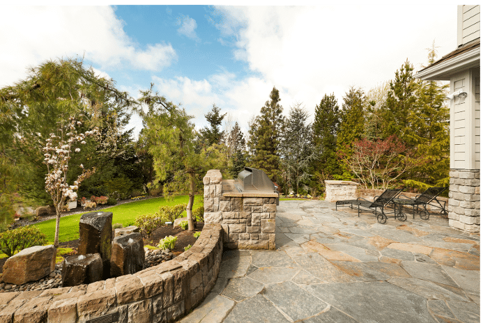 A stone patio with lounge chairs, a built-in grill, and a landscaped garden featuring a small stone waterfall and trees, set against a backdrop of a blue sky with scattered clouds.