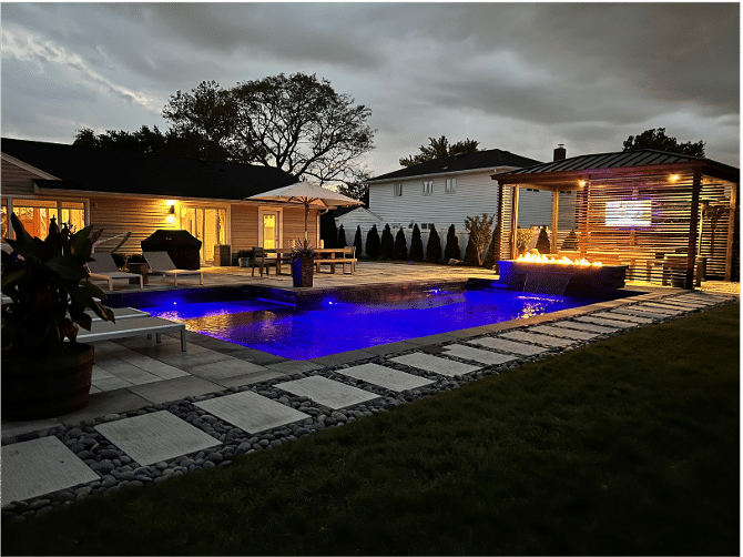 A modern backyard features a lit swimming pool surrounded by stone tiles, lounge chairs, a pergola with a TV, and a fire pit under a cloudy evening sky.