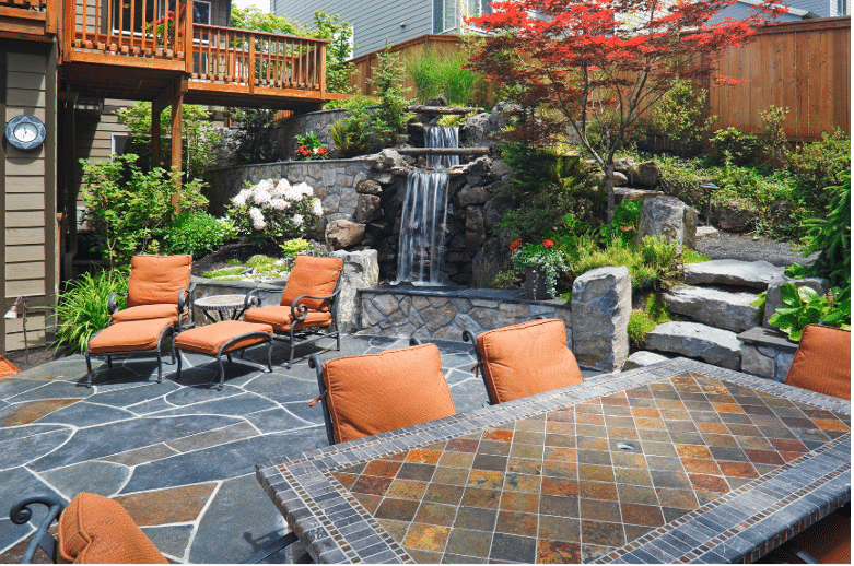 A landscaped backyard with a stone patio, orange cushioned chairs, a tiled table, a small waterfall, and lush greenery.