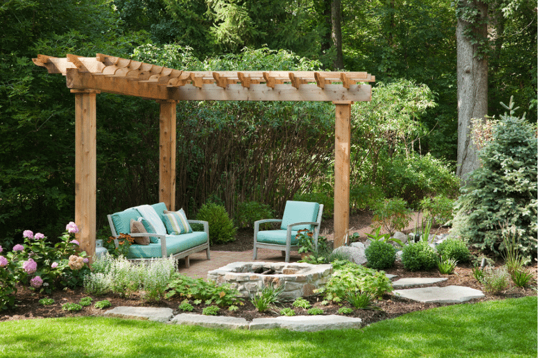 A garden with a wooden pergola, blue cushioned chairs, a stone fire pit, and surrounding greenery and flowers on a sunny day.