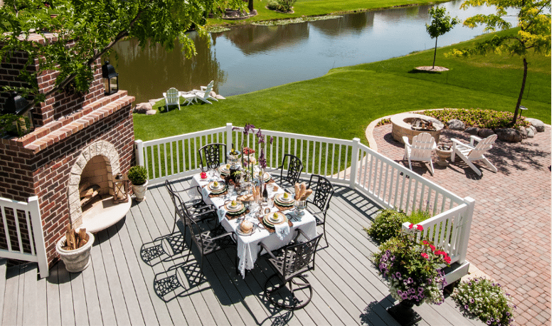 An outdoor deck features a dining table set for a meal next to a brick fireplace, with views of a pond, green lawn, and a small patio area with Adirondack chairs nearby.