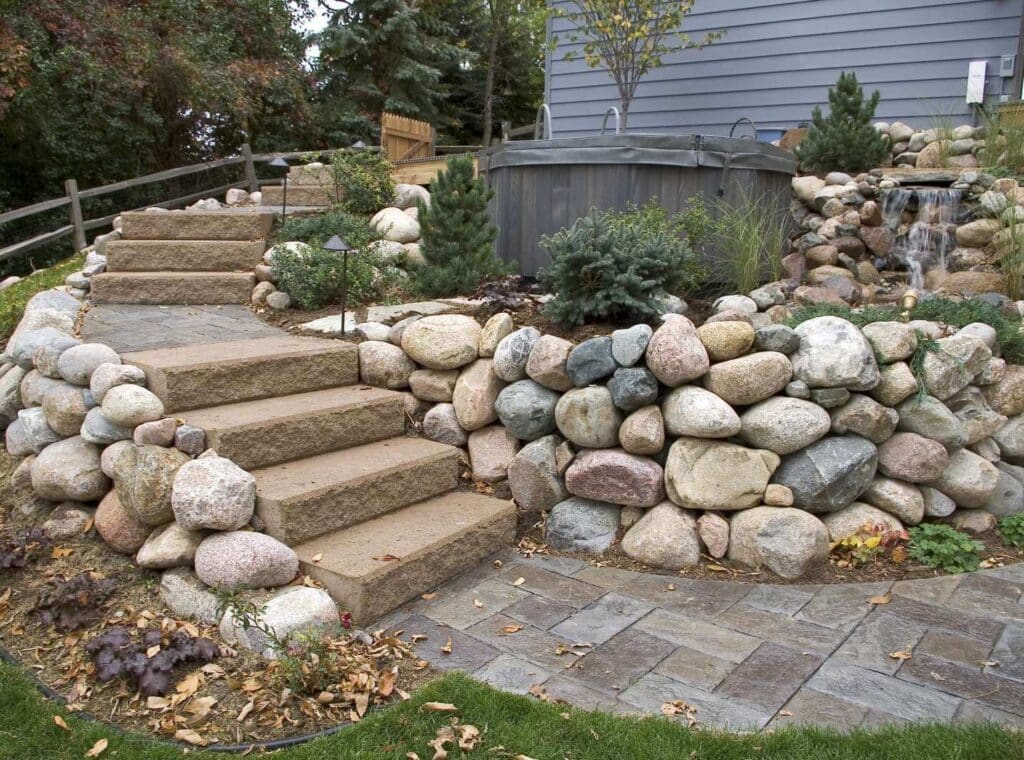 Stone steps lead to a hot tub surrounded by a rock wall and landscaping with small trees and shrubs, next to a paved walkway.