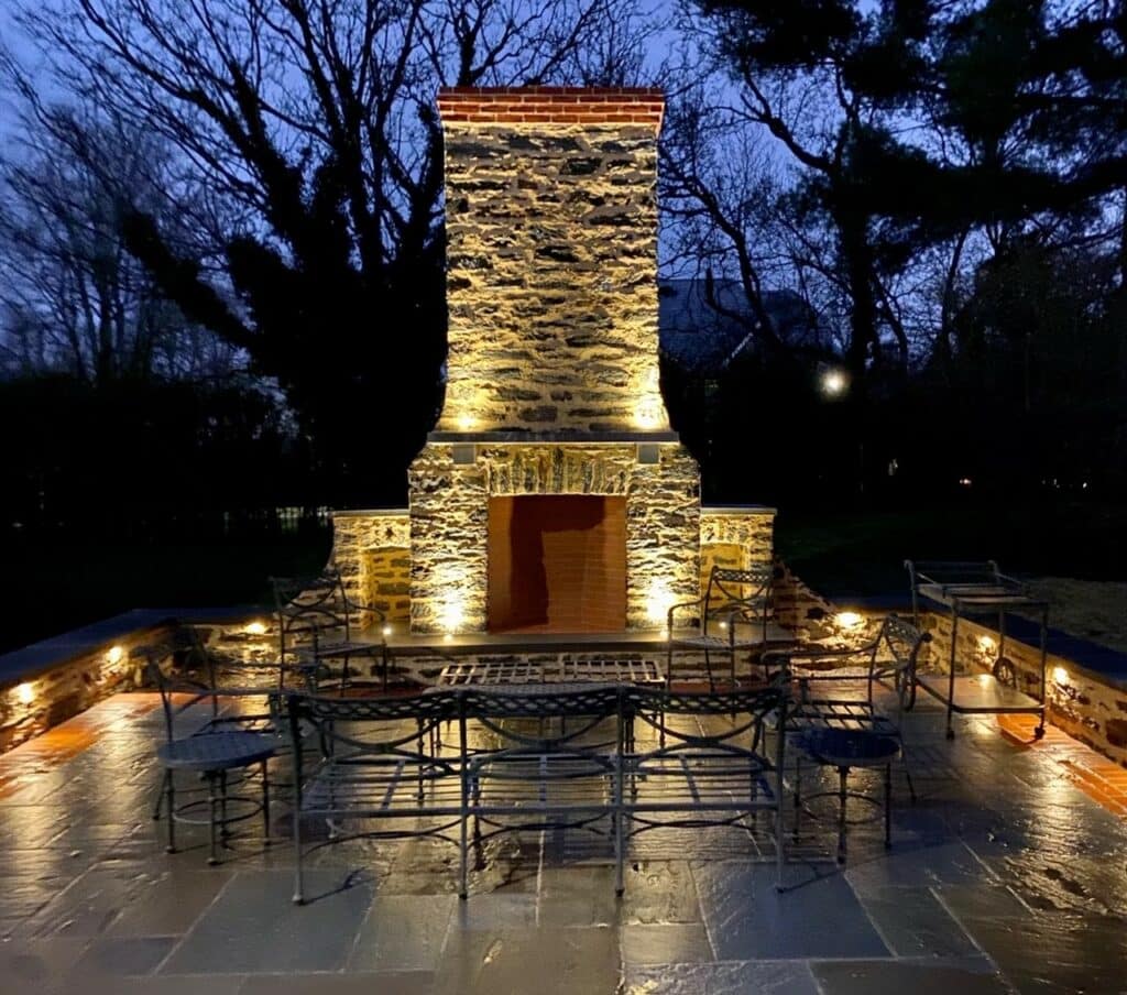 Outdoor stone fireplace illuminated at night, surrounded by metal chairs on a stone patio, with trees in the background.