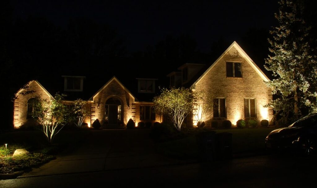 A large brick house at night illuminated by outdoor lights, with trees and shrubs along the driveway.