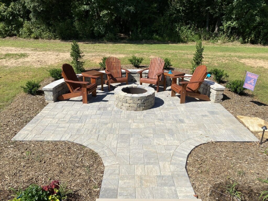 A beautifully crafted stone patio with five wooden chairs encircles a circular stone fire pit. This serene setting, enhanced by expert landscape design, is surrounded by lush greenery with a small red flag in the background.