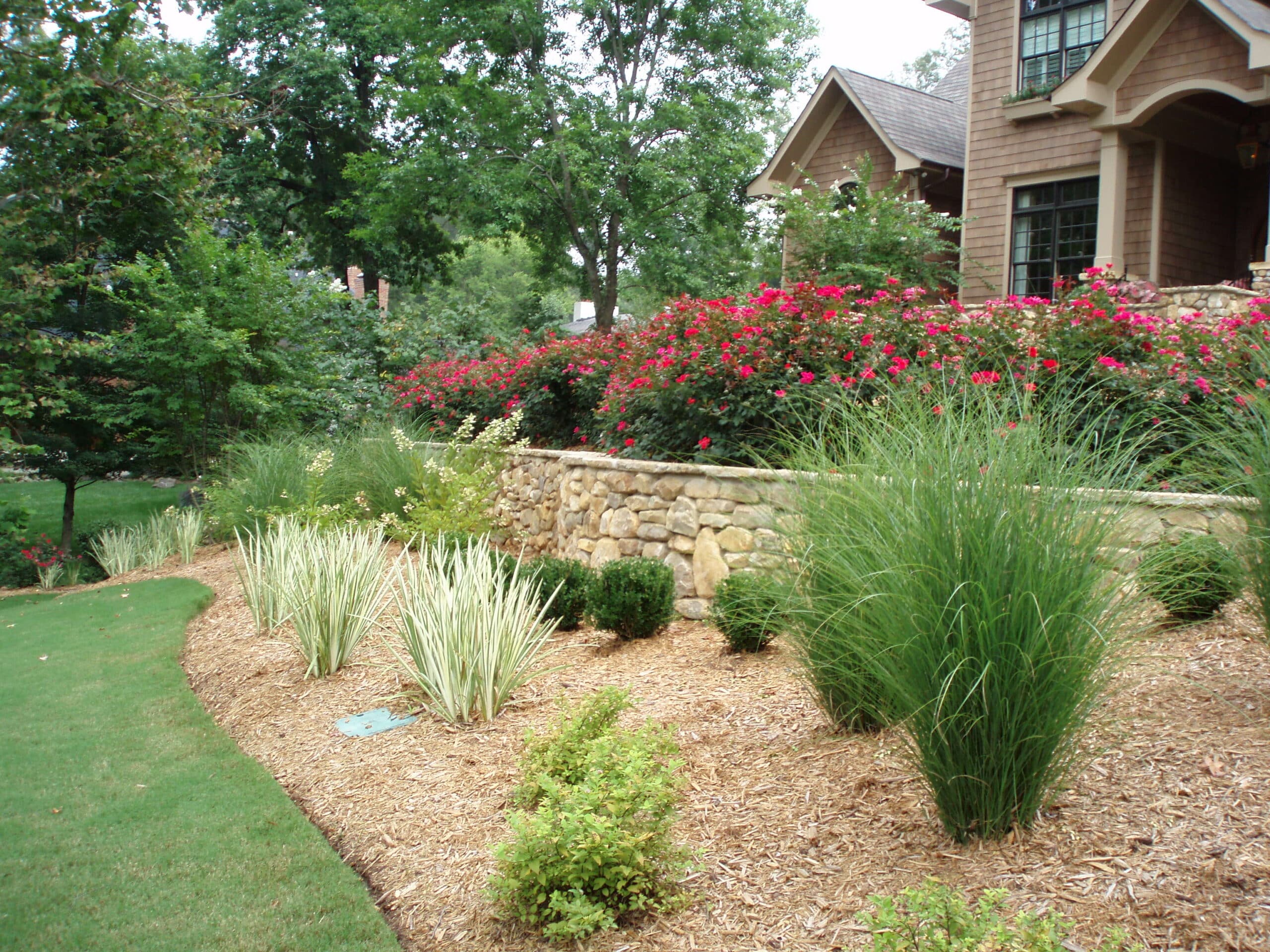 Well-maintained garden with green grass, ornamental grasses, and flowering shrubs in front of a brick house. Stone retaining wall separates lawn from garden beds. Trees and plants in background.