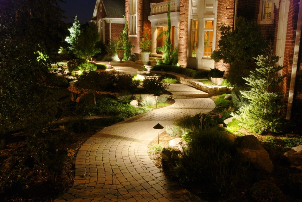 A brick pathway winds through a garden with plants and shrubs, leading to a well-lit brick house entrance at night.