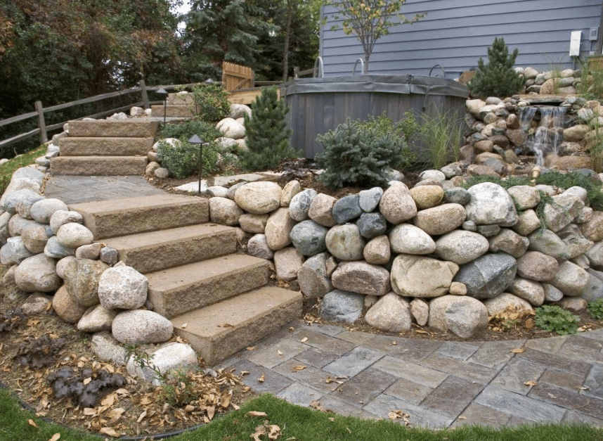 Outdoor garden area with stone steps leading to a raised hot tub. Surrounded by large rocks, small trees, and a stone pathway.