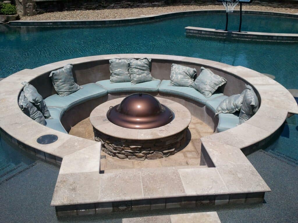 Circular recessed seating area with a fire pit in the center, surrounded by a large swimming pool. Expert landscaping design complements the cushioned seats that encircle the fire pit, creating a serene retreat reminiscent of top-notch spaces found in Charlotte, NC.