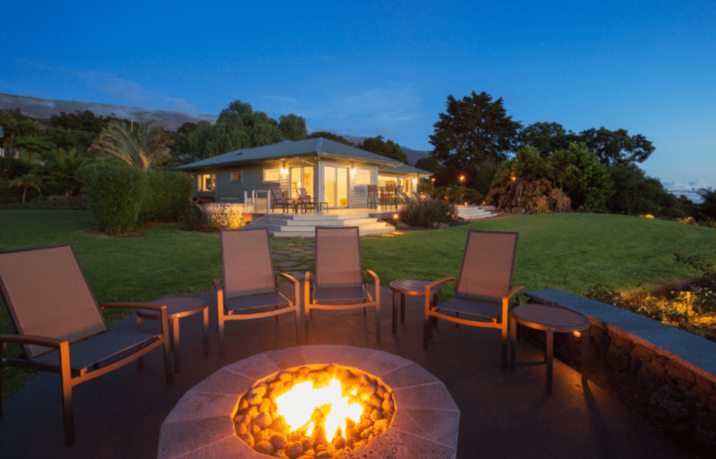 A cozy, outdoor fire pit surrounded by chairs on a patio. In the background, a modern house with a well-lit interior and a spacious lawn is visible at dusk.