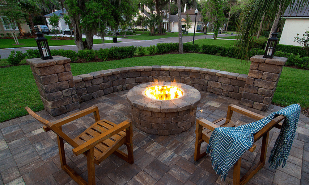 A stone fire pit with a lit fire is surrounded by two wooden chairs on a brick patio, with a blue blanket draped over one chair. Trees and houses are visible in the background.