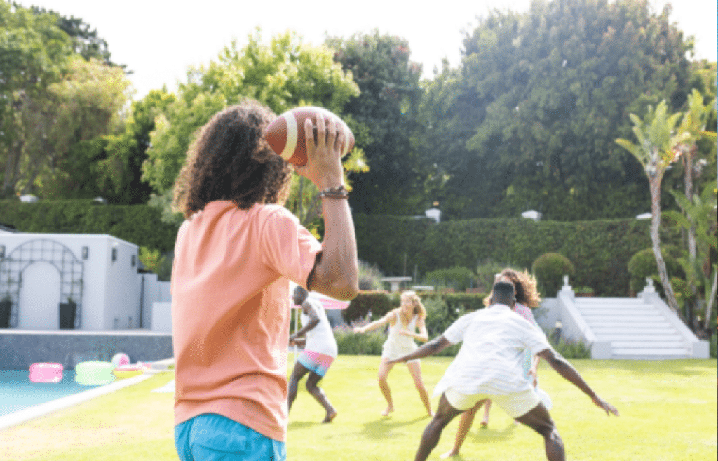 People are playing football in a grassy backyard near a pool on a sunny day.