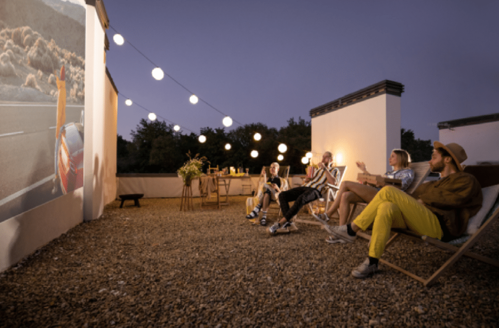 A group of people sit on lounge chairs watching a film on an outdoor projector screen in a cozy, decorated rooftop setting at dusk.