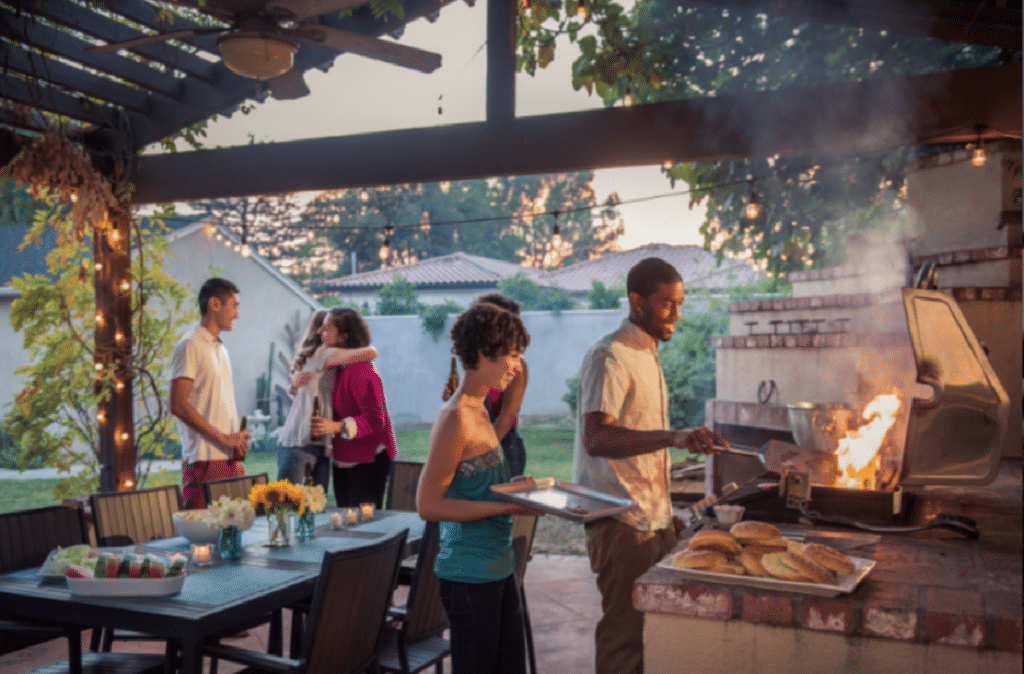 People enjoy a backyard barbecue gathering. A person cooks on a grill while others socialize nearby. A table is set with flowers and food under a pergola.