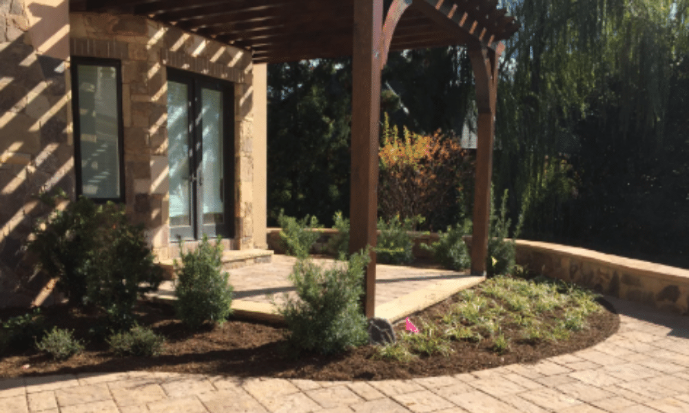 A stone patio with a wooden pergola, surrounded by freshly planted shrubs and a small garden bed, adjacent to a house.