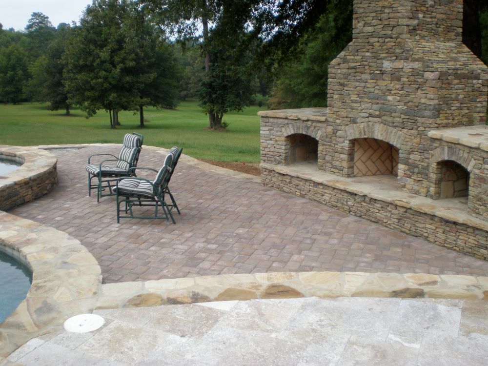 Stone patio with built-in fireplace, three striped chairs, and a pool edge in a grassy outdoor setting.