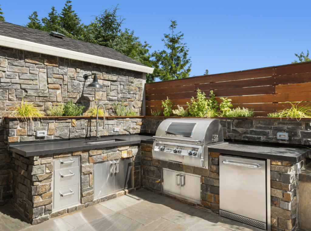 Outdoor kitchen with stone walls, stainless steel appliances, and a grill. Countertop with sink and planted herbs. Wooden privacy fence and trees in the background under clear blue sky.