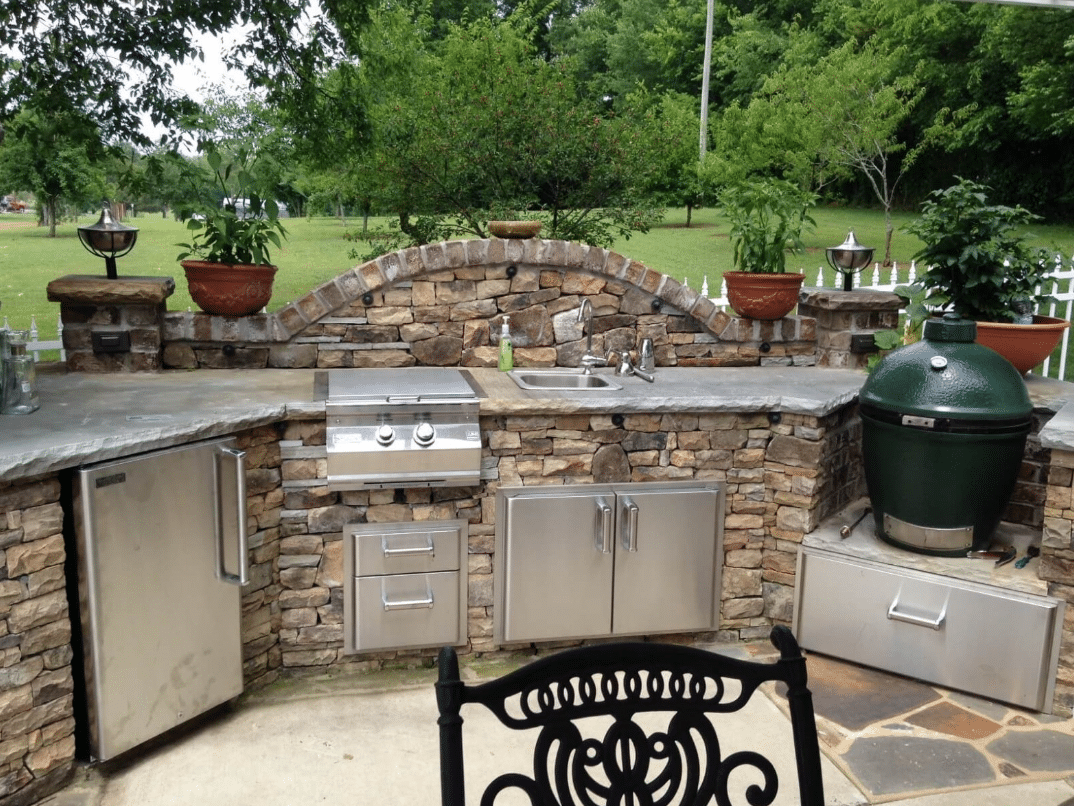 Outdoor kitchen with stone countertops, built-in grill, smoker, sink, mini-fridge, and decorative pots, set against a green lawn and trees.