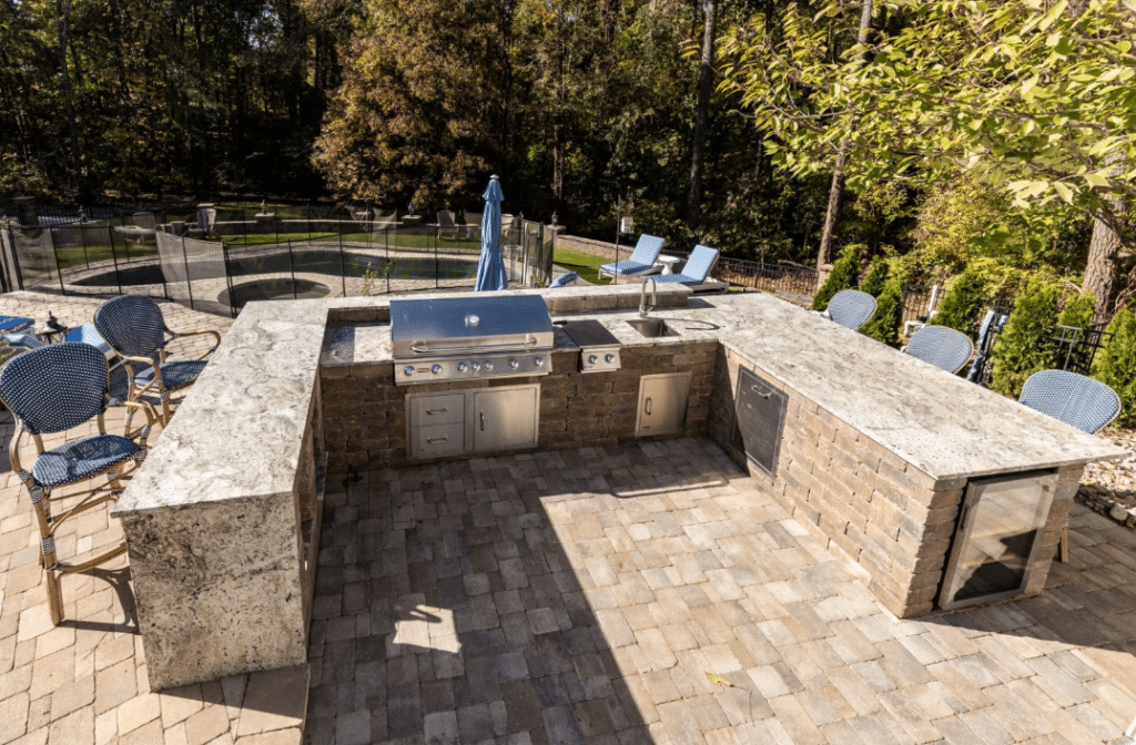 Outdoor kitchen with stone countertops, grill, and sink. Patio chairs surround the setup. Trees and a pool are visible in the background.