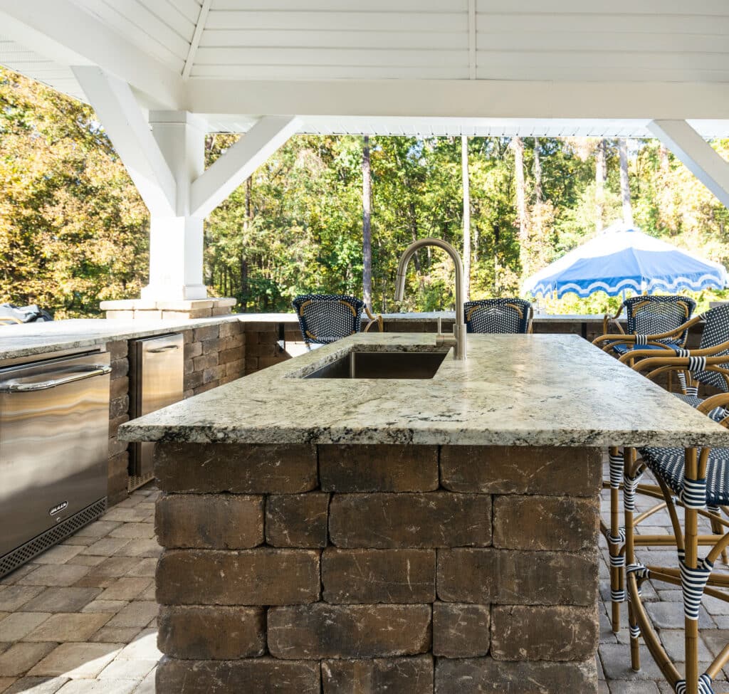 Outdoor kitchen with a stone island featuring a sink, surrounded by bar stools, and a stainless steel grill under a white pergola. An umbrella and trees are visible in the background.