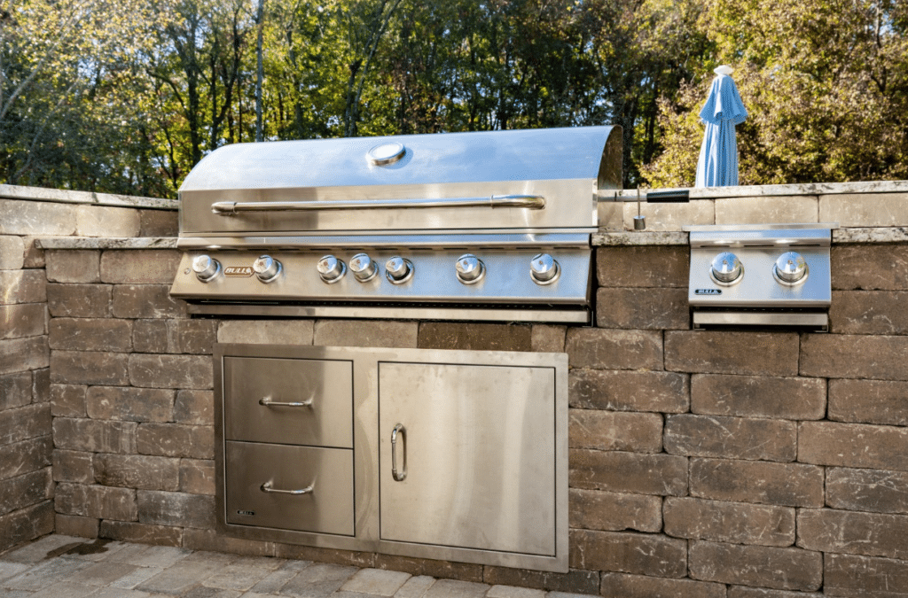 Stainless steel outdoor grill with multiple knobs, set into a brick countertop. Trees and a blue umbrella are visible in the background.