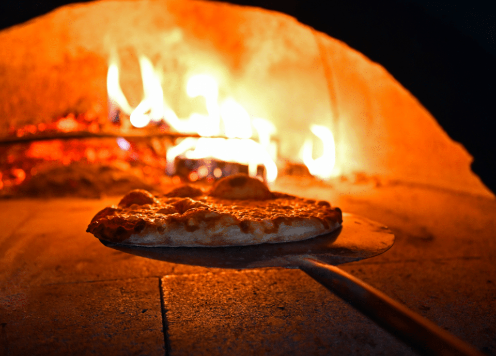 Pizza being cooked on a peel inside a wood-fired oven with visible flames.