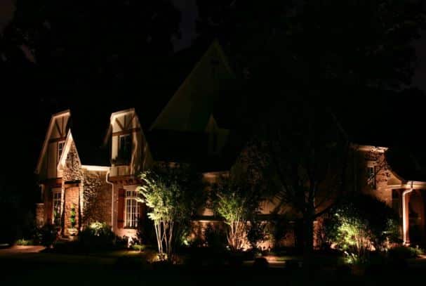 A large house with peaked roofs is illuminated by outdoor lights at night, surrounded by trees and shrubs.