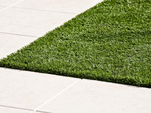 A section of green artificial grass bordered by light-colored square tiles in an outdoor setting.