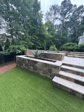 Stone steps lead to a raised patio with a built-in planter containing a large plant; artificial grass covers the lower level, bordered by trees and a black metal fence.