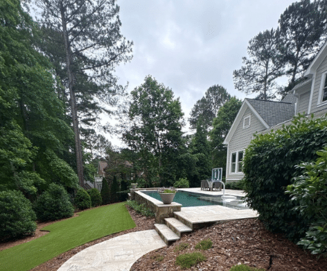 Stone steps lead to a backyard pool beside a gray house, surrounded by trees, shrubs, and landscaping under an overcast sky.