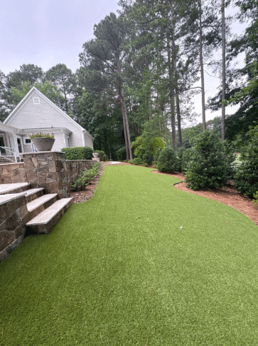 A backyard with artificial turf, bordered by shrubs and trees, stone steps on the left, and a white house in the background.