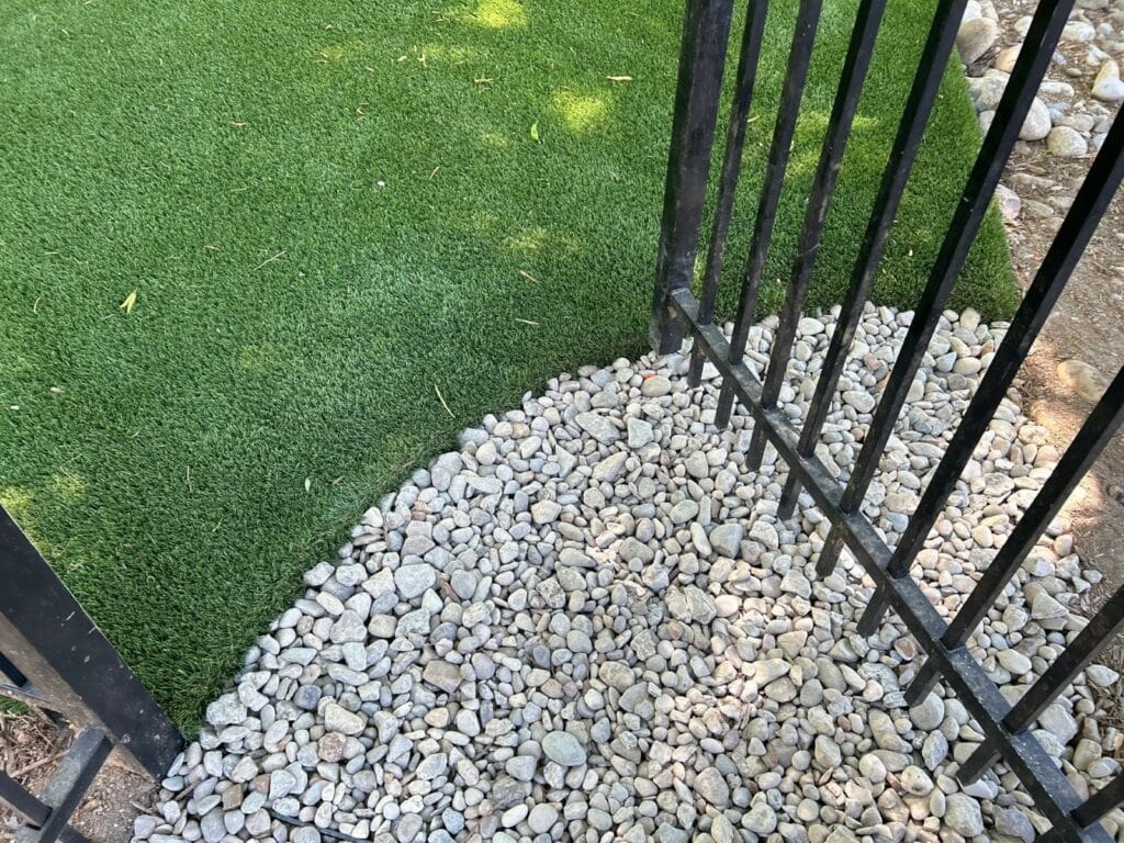 A black metal fence separates an area of artificial grass from a section covered with small white rocks.