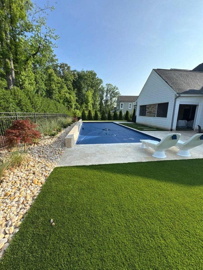 A backyard with artificial grass, a stone path, two white lounge chairs, and a covered swimming pool next to a white house. Shrubs and trees line the background.