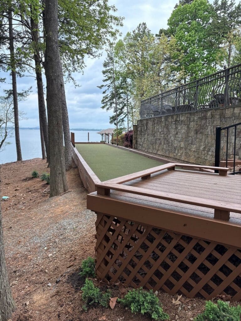A bocce ball court with artificial turf sits beside a stone retaining wall, surrounded by trees and overlooking a lake under a cloudy sky.