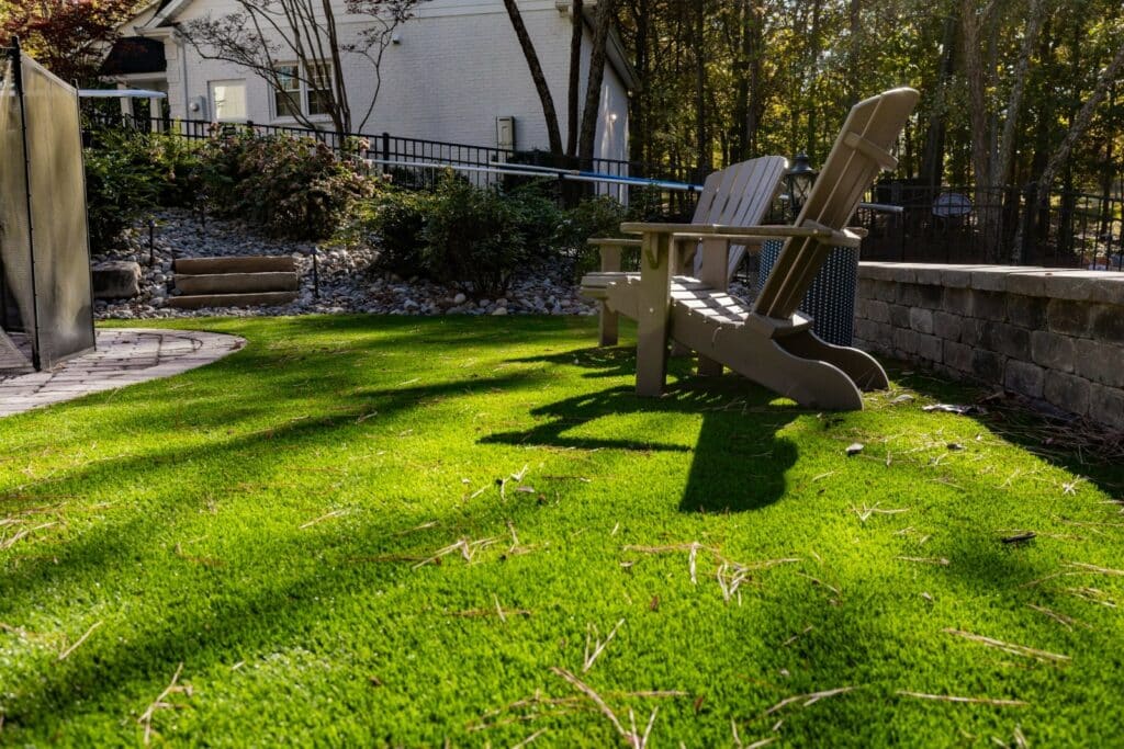 Two Adirondack chairs sit on a lawn near a stone patio, with a white house, shrubs, and trees visible in the background. Sunlight casts shadows on the grass.
