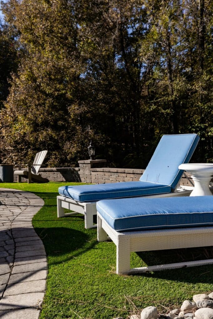 Two white outdoor lounge chairs with blue cushions sit on artificial grass beside a curved stone pathway, with trees and patio walls in the background.