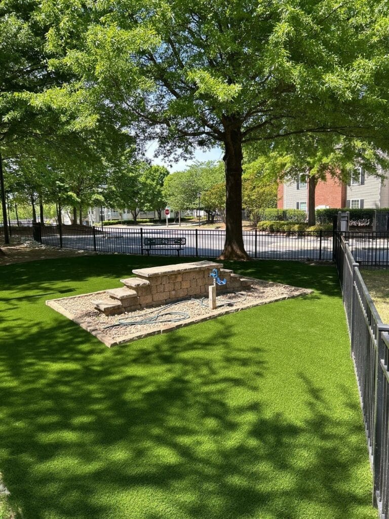 A small fenced area with artificial grass, a tree providing shade, a stone planter, a blue hydrant, and nearby residential buildings in the background.