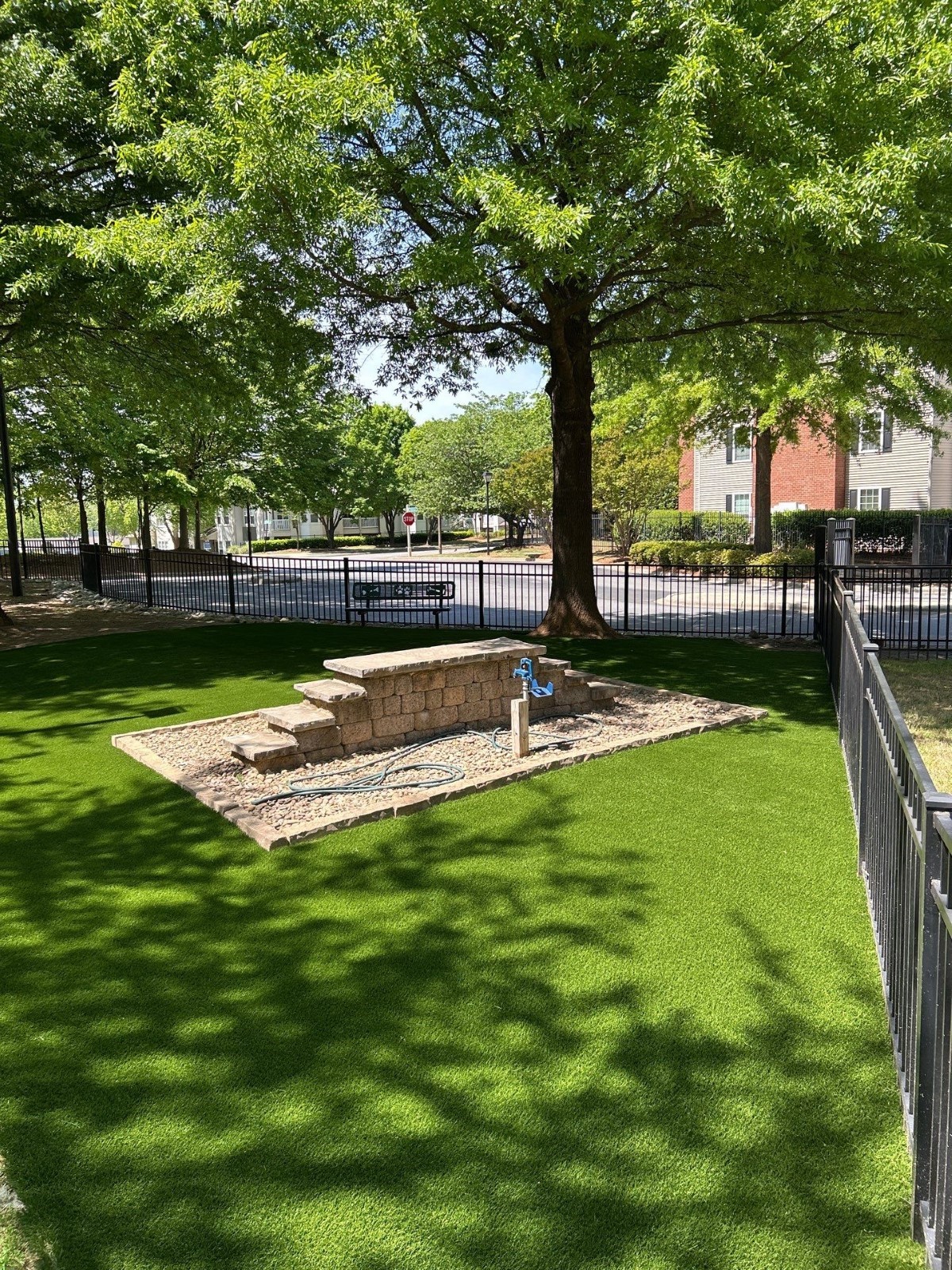 A small fenced area with artificial grass, a tree providing shade, a stone planter, a blue hydrant, and nearby residential buildings in the background.